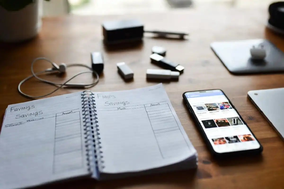 Notebook with savings plan next to gadgets and a phone showing deals