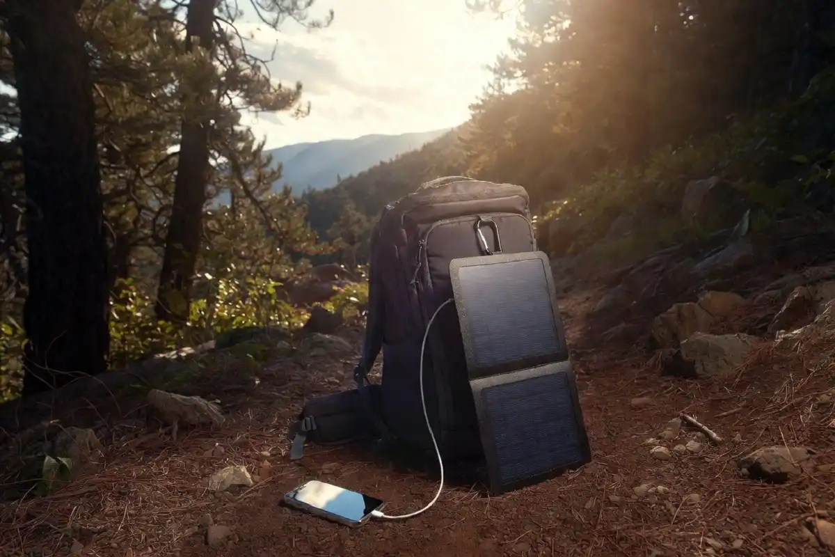 A foldable solar panel charger clipped to a backpack charging a smartphone on a sunny hiking trail surrounded by trees