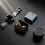 A flat lay of smart gadgets for men including a smartwatch, wireless earbuds, and compact power bank on a dark desk