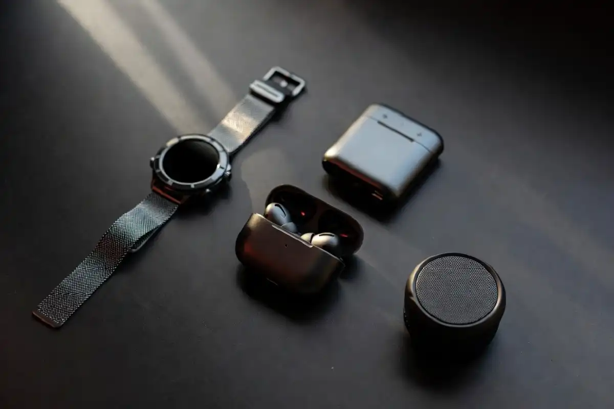 A flat lay of smart gadgets for men including a smartwatch, wireless earbuds, and compact power bank on a dark desk