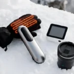 A flat-lay collection of popular winter gadgets including heated gloves, electric hand warmer, smart thermostat, and heated travel mug on a snowy white surface