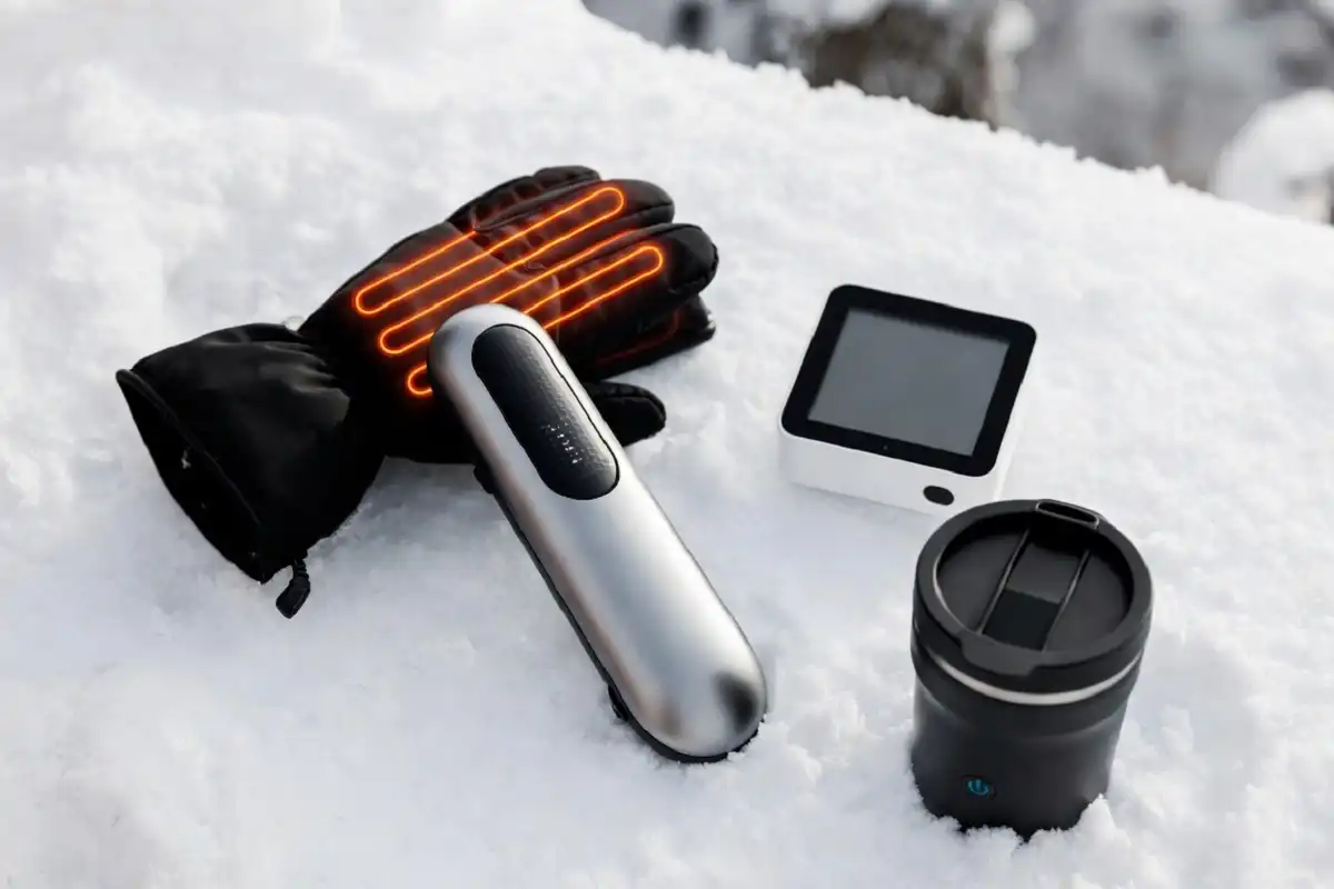 A flat-lay collection of popular winter gadgets including heated gloves, electric hand warmer, smart thermostat, and heated travel mug on a snowy white surface