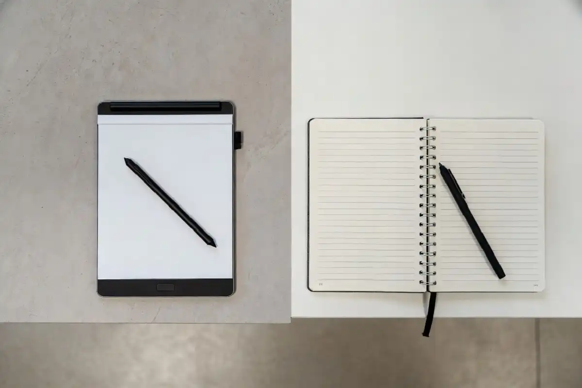 Electronic paper note tablet placed next to an open paper notebook and pen for comparison